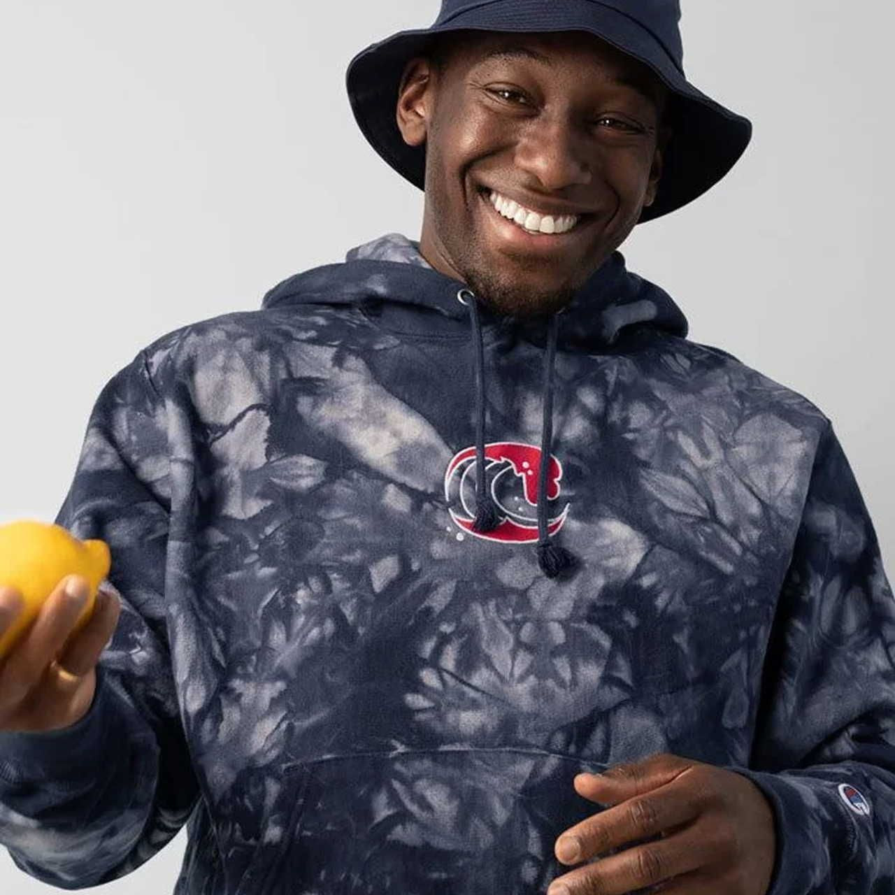 A smiling man wearing a navy tie-dye hoodie with a logo holds a lemon and wears a navy bucket hat.